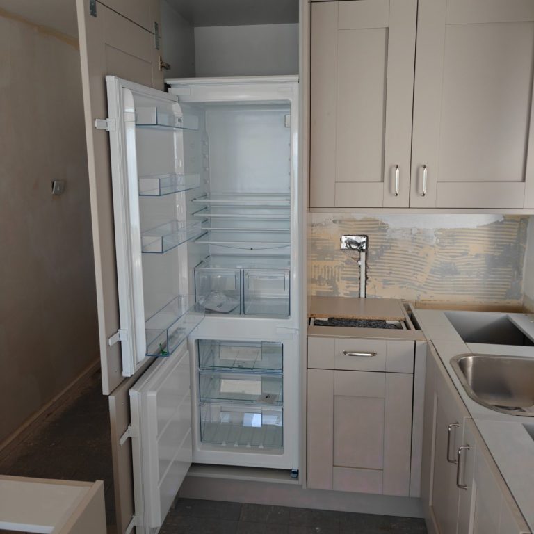 Waiting for worktops Empty kitchen fridge with an open door, featuring a grey cabinet and sink.